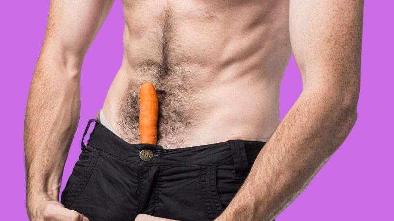 A photo of a man displaying penile girth enlargement techniques with a carrot in his pants.